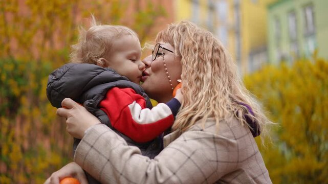 Slow motion. Close-up. A little son hands a tangerine to his mother, while standing next to her on the grass in the park on a spring day. The mother smiles hugging the child and kisses him on the lips