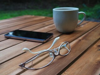 木製テーブルの上に置かれているコーヒーカップとスマホとメガネ