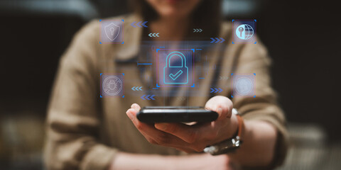 A woman holding a smartphone with digital cybersecurity icons hovers on screen as a virtual...