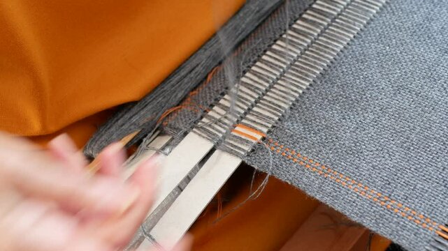 Hands make stitches with a needle and gray thread along the edge of a woven product, selective focus. Completing a woven project