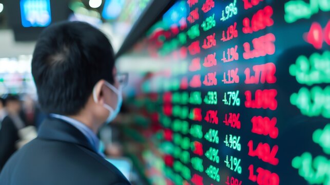 A man wearing a mask closely observes stock market trends displayed on a screen, focusing on financial data and market fluctuations.