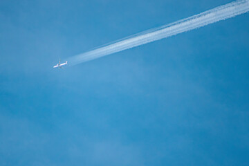 An airplane flies high in a clear blue sky, leaving a long contrail behind. The scene captures the vastness of the sky and the speed of travel. Ample copy space.