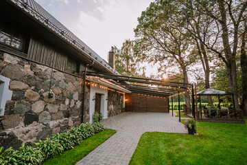 Blome, Latvia - September 11, 2023 - Charming stone and wood house with a pergola and garden at sunset, featuring warm lighting and lush greenery. Copy space...