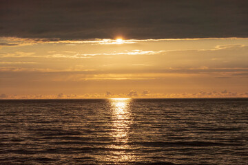 Sunset over the sea. There is a clear bright orange strip of sky between the dark surface of the water and the clouds. The sun's path is visible on the surface of the sea.