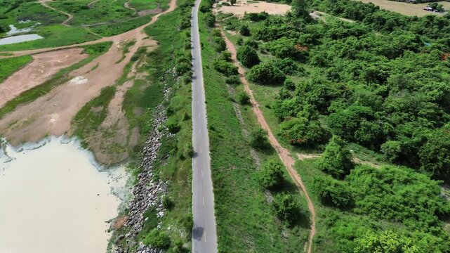Drohnenaufnahme zeigt eine lange, gerade Stra&szlig;e, die durch eine &uuml;ppige, gr&uuml;ne Landschaft in Thailand verl&auml;uft. Links der Stra&szlig;e befinden sich sandige Fl&auml;chen und ein Gew&auml;sser, rechts dichter Wald.