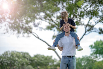 Fototapeta premium White son sitting on his father shoulder with smile and fun, looking his father with love walking in park with blur background and big tree and copy space, relationship between dad and son.