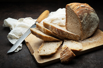 Traditional bread cut into slices. Loaf of bread with knife and cutting board on dark background.