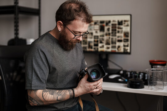 Focused photographer with tattoos inspects his camera lens in a well-organized workspace with photography equipment. - Powered by Adobe