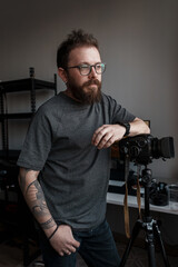 Confident male photographer with beard standing in a studio, posing thoughtfully next to his camera mounted on a tripod.