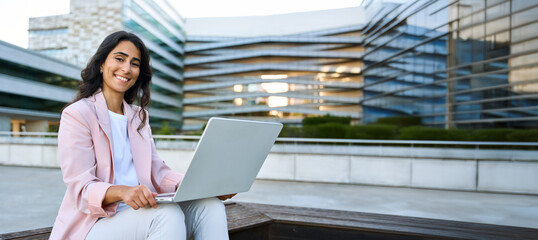 Banner with smiling portrait of young middle eastern Israel businesswoman using laptop pc. Indian or Arabian business woman freelancer in suit working online remotely on computer, outdoors. Copy space