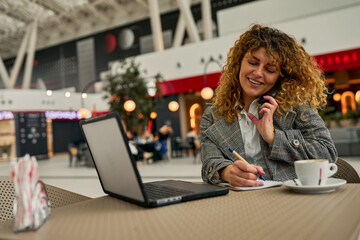 Successful and cheerful, a caucasian businesswoman takes notes, multitasks with her laptop, and sips coffee at the train station cafe.