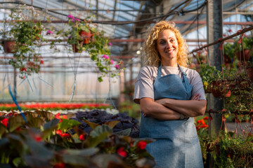 Confident woman standing arms crossed in her family’s greenhouse surrounded by lush plants.