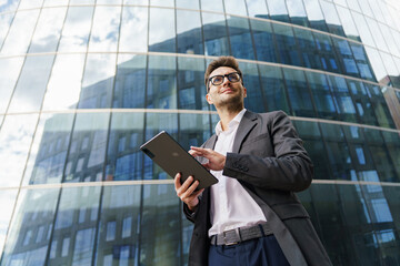 A confident businessman in a suit holds a tablet, standing against a backdrop of modern glass buildings.