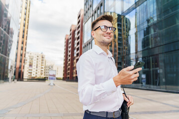 A young businessman, smiling confidently, holds his phone in a modern urban setting.