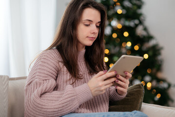 Woman in a cozy sweater using a tablet by the Christmas tree at home, relaxed holiday atmosphere.