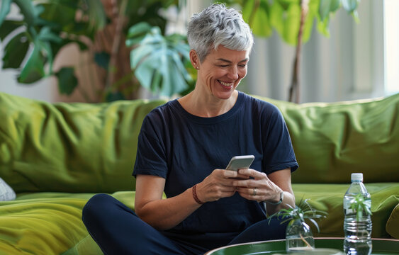 A middle-aged woman in dark blue yoga pants sits on the floor of her living room, smiling at an iPhone she is holding and reading. She has short silver hair tied back with some graying strands