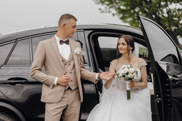 A man and a woman are getting married and are standing in front of a black car. The man is wearing a suit and tie, and the woman is wearing a white dress. They are both smiling and seem happy