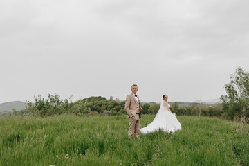 A bride and groom stand in a field of grass. The bride is wearing a white dress and the groom is wearing a suit. The sky is cloudy and the grass is lush and green. Scene is romantic and peaceful