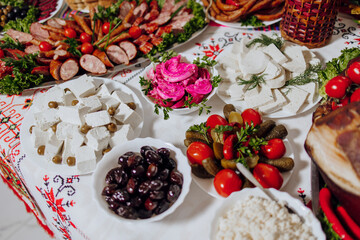 A table is covered with a variety of foods, including cheese, olives, tomatoes, and pickles. The tablecloth is embroidered with a floral pattern