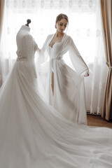 Naklejka premium A woman stands in front of a white gown, which is displayed on a mannequin. The gown is long and flowing, and the woman is wearing a necklace. Concept of elegance and sophistication