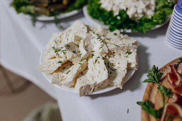 A plate of cheese and herbs sits on a table with other plates of food. The cheese is cut into small cubes and is garnished with parsley