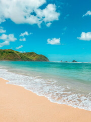 Obraz premium A pristine tropical beach in Lombok with golden sand, clear turquoise water, and lush green hills under a bright blue sky with scattered clouds, showcasing the serene beauty of this paradise. 