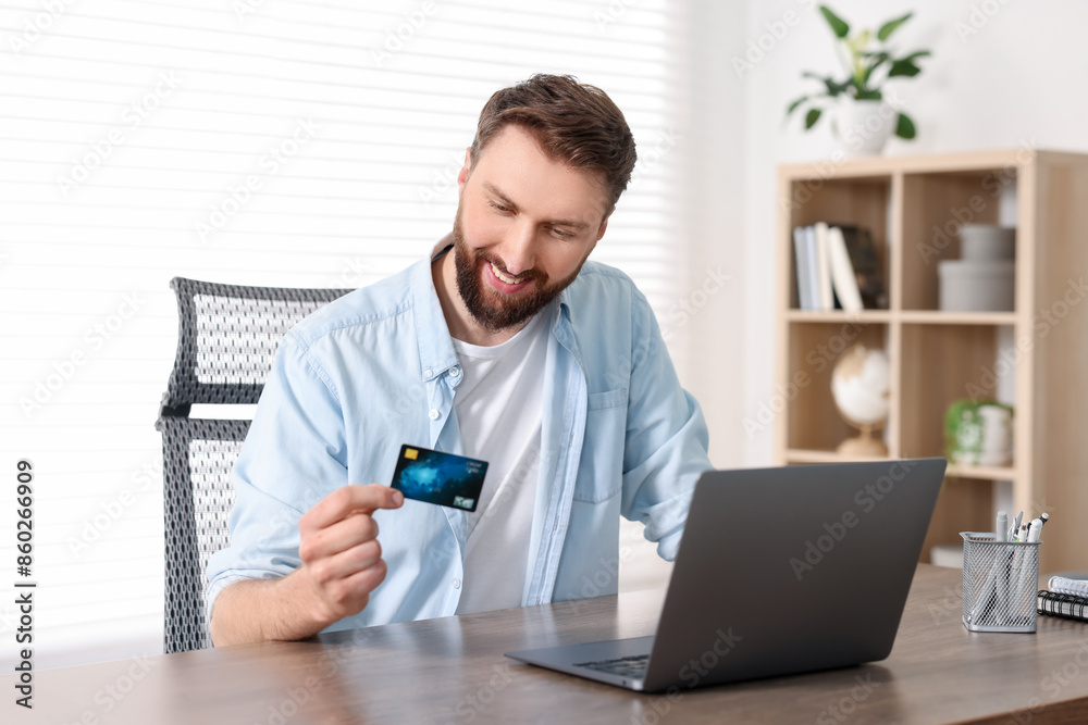 Wall mural Online banking. Happy young man with credit card and laptop paying purchase at table indoors