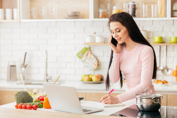 A young woman is multitasking in her modern kitchen. She is talking on the phone while taking notes, surrounded by fresh vegetables, a laptop, and a pot on the stove.