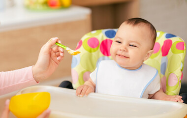 A mom is feeding her baby boy who is sitting in a high chair in the kitchen. The scene is warm and homey, emphasizing the nurturing bond during mealtime.