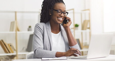 Smiling young African American businesswoman in formal dress, working on a laptop in the office, on a phone call.