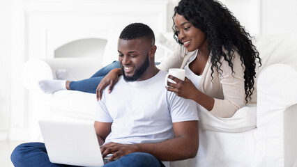 Fototapeta premium A couple relaxes on a white couch, one working on a laptop while the other holds a mug of coffee.