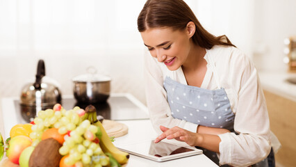 A woman wearing an apron stands in her kitchen and uses a tablet to find a recipe.
