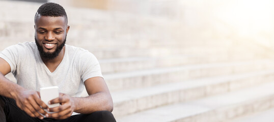 A man with a beard sits on a set of steps, looking down at his phone with a smile. © Prostock-studio