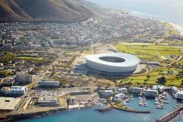 Fototapeta premium Harbor, drone and stadium in city for travel with ships, cruise or buildings by ocean and mountain. Boats, coast and aerial view of infrastructure by seashore for holiday destination in Cape Town.