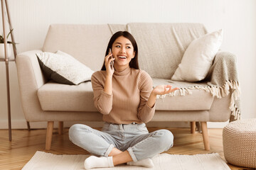 Obraz premium A young woman sits on the floor of a living room, smiling while talking on her phone.