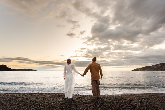 A couple standing on a beach holding hands. The sky is cloudy and the sun is setting. Sceane is romantic and peaceful