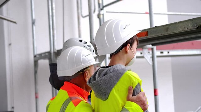 experienced male builder in yellow protective jacket, children in white hardhats came on excursion, father, grandfather tells grandchildren about his profession with background of high buildings