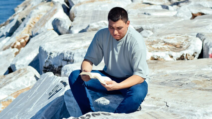 Young man reading a book sitting on the rocks on the beach