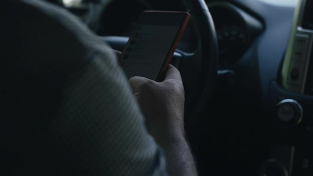 A close-up shot of a drivers hand holding a smartphone while driving on a street.