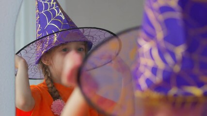 A little girl of 5 years old in an orange T-shirt with disheveled hair stands in front of a mirror dressing up for Halloween.