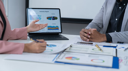 A photograph of a male and female employee's hands as they meet in an office, pens and laptops at the ready as they consult on business matters.