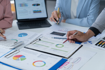 A photograph of a male and female employee's hands as they meet in an office, pens and laptops at the ready as they consult on business matters.
