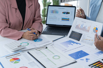 A photograph of a male and female employee's hands as they meet in an office, pens and laptops at the ready as they consult on business matters.