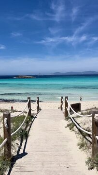 Vertical video. White sand beach on a narrow strip of land on Formentera. Blue famous beach with people relaxing on vacation in the mediterranean sea. Turquoise sea. Beach with a wooden bridge.