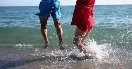 Happy man and woman jump high on beach edge washed by sea waves on sunny day. Couple in love enjoys summer vacation at resort slow motion