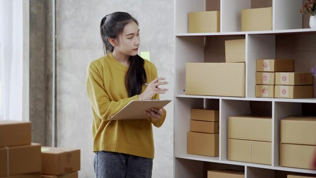 Asian woman managing inventory in a warehouse storage room. Checking and marking boxes, organizing products on shelves with a clipboard