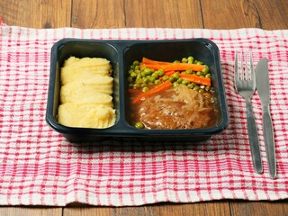 A meal consisting of burger, mashed potatoes and vegetables in plastic tray is served on a red and white checkered tablecloth and wooden table. Simple classic dish for take away. Office lunch.