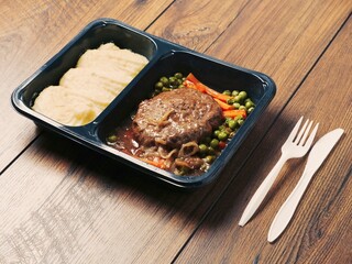 A meal consisting of burger, mashed potatoes and vegetables in plastic tray is served on wooden table and plastic fork and knife. Simple classic dish for take away. Office lunch.