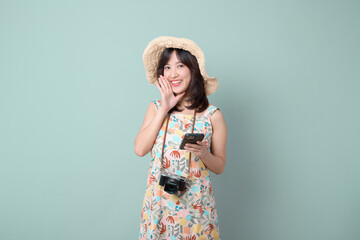 Travel and vacation promotion concept. Happy Asian woman wearing casual dress and hat with camera and mobile phone while shouting to free copy space isolated on pastel green background.