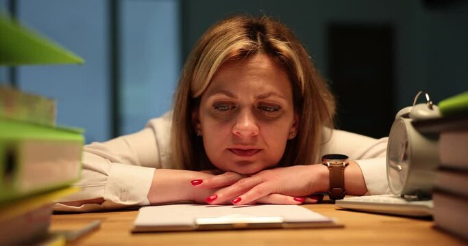 Pensive woman leans on wooden table near clipboard thinking about vacation while lightning flashing. Employee stays late at work in office feeling tired